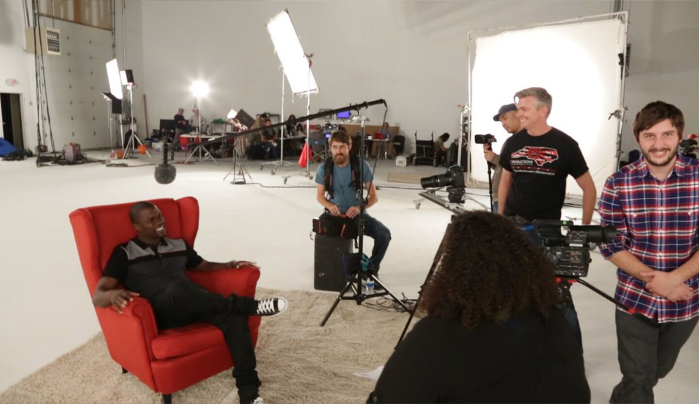 Corporate video production shoot in white studio, man in red chair being filmed by crew