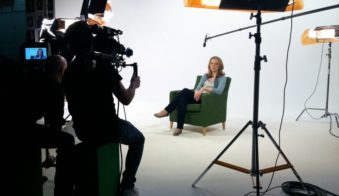 Corporate interview video shoot in white studio, woman in green chair filmed by camera crew