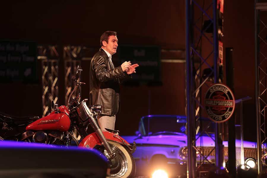 Speaker presenting on stage beside a vintage Harley-Davidson motorcycle at a corporate conference event