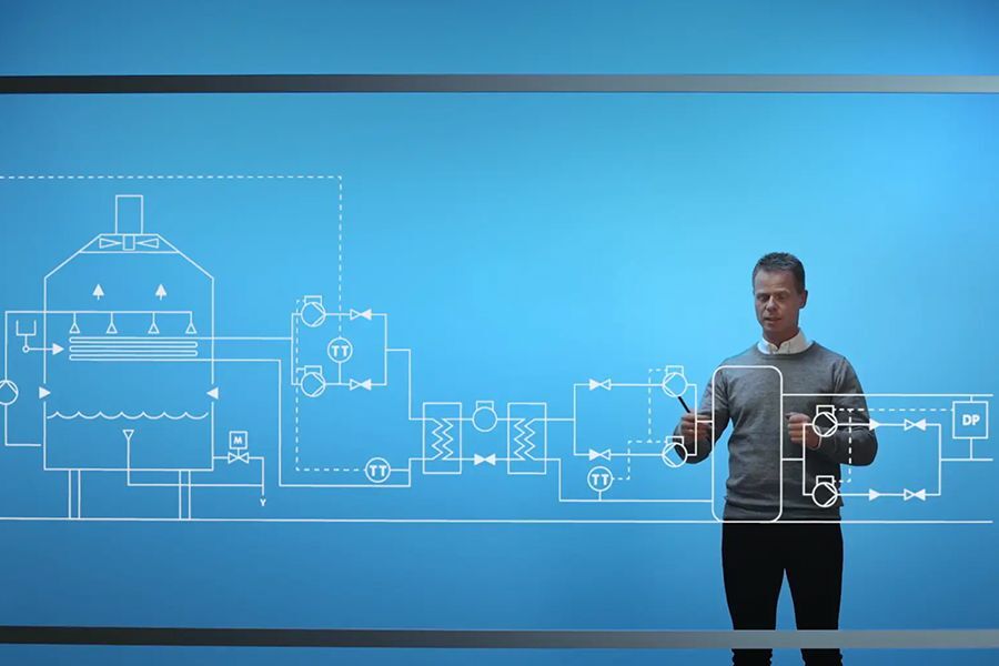 Grundfos presenter standing before a large blue LED display with technical piping diagrams during a corporate webcast